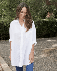 A woman with long brown hair smiles while looking down, wearing the Mackenzie in White Linen shirt by Frank & Eileen and blue jeans. She stands outdoors on a gravel path with greenery in the background.