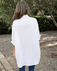 A person with long brown hair stands outdoors on a gravel path, surrounded by greenery, wearing Frank & Eileen's Mackenzie in White Linen shirt and blue pants.