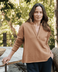 A woman with long brown hair wears a Frank & Eileen Patrick Henley in Camel Terry and dark pants as she stands by a wooden table outdoors, smiling softly amid lush greenery and sunlight.