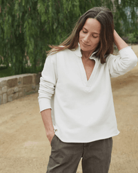 A woman with long brown hair stands outdoors, smiling slightly as she wears the Frank & Eileen Patrick Henley in Vintage White Terry and gray pants, one hand in her pocket and the other touching her hair, framed by greenery and a stone wall.