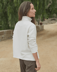 A woman with long brown hair stands outdoors on a dirt path, wearing the Frank & Eileen Patrick Henley in Vintage White Terry and dark pants. She faces away, framed by greenery and a stone wall.
