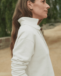 A woman with long brown hair stands outdoors in a Frank & Eileen Patrick Henley in Vintage White Terry, sleeves pushed up. She’s shown in profile with greenery and a path behind her.