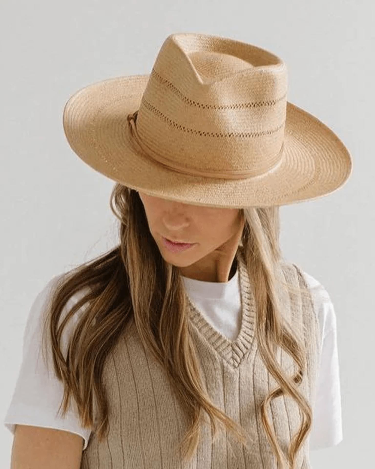 A person with long, wavy hair looks downward against a plain light background, wearing the Gigi Pip Arlo Honey Straw Teardrop Fedora with Nude Band, styled with a white t-shirt and beige knit vest.