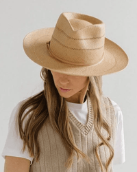 A person with long, wavy hair looks downward against a plain light background, wearing the Gigi Pip Arlo Honey Straw Teardrop Fedora with Nude Band, styled with a white t-shirt and beige knit vest.