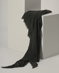 The Grisal Love Cashmere Scarf in Charcoal is draped casually over two minimalist white blocks, its luxurious fabric cascading down and forming gentle folds and shadows against a plain background.