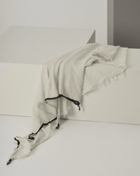 The Grisal Rosa Cashmere Scarf in Milk X Black, featuring a black trim and frayed edges, is draped over a white geometric block against a minimalist neutral background. Its light, semi-transparent texture adds effortless style.