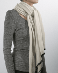A person wears a long-sleeved, gray ribbed shirt and the Grisal Rosa Cashmere Scarf in Milk X Black wrapped around their neck, standing against a plain, light background.