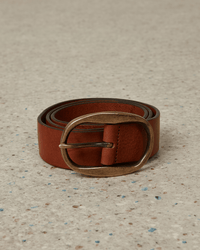 A rolled-up Hartford Angus Leather Belt in Brown, featuring a large oval brass buckle, rests on a speckled light surface.