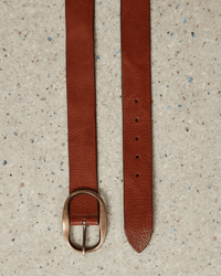 The Hartford Angus Leather Belt in Brown, made of genuine leather with a round brass buckle, is displayed on a light speckled surface. Its textured brown leather and five adjustment holes add both style and function.
