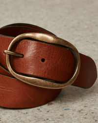 A close-up of the Angus Leather Belt in Brown by Hartford, featuring an antique brass oval buckle, coiled on a speckled light-colored surface.