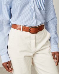Person in a light blue striped shirt tucked into high-waisted cream pants, accessorized with the Hartford Angus Leather Belt in Brown—a wide leather belt with a large buckle. Only their torso and upper legs are visible.