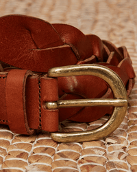 A close-up of the Hartford Arthur Leather Belt in brown, showcasing its braided genuine leather design and brass buckle, displayed on a woven surface.