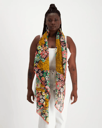 A woman in a white outfit stands against a plain background, wearing Inoui Editions' Scarf 100 Pampa in Multicolor—crafted from 100% cotton with vibrant floral and leopard prints—draped around her neck.