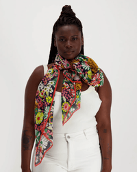 A woman in a white sleeveless top and pants faces forward, wearing the Inoui Editions Scarf 100 Pampa in Multicolor wrapped around her neck. She has long braids, visible arm tattoos, and her scarf is made from 100% cotton.