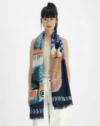 A woman with straight black hair and bangs stands against a plain background, wearing a sleeveless beige top, white pants, and an Inoui Editions Scarf 70 Toucan in Emerald with blue, orange, and green patterns and fringed edges.