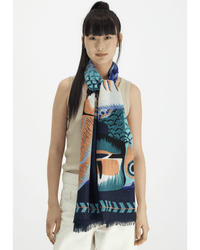 A woman with long dark hair and bangs wears a sleeveless beige top, off-white pants, and the Inoui Editions Scarf 70 Toucan in Emerald, featuring vibrant blue, orange, and green tones, set against a plain white background.