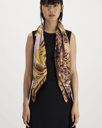 A woman with straight dark hair and bangs wears a sleeveless black dress and drapes the Inoui Editions Square 100 Chatou in Pink scarf around her neck, standing against a plain white background.