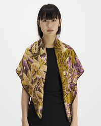 A woman with straight black hair and bangs wears a black top and the Inoui Editions Square 100 Chatou in Pink, an oversized scarf featuring a bold, colorful floral and tiger print against a plain white background.