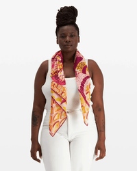 A woman with dark skin and braided hair in a bun wears a white sleeveless top and pants, accessorized with the Inoui Editions Square 100 Tango in Yellow bandana—100% cotton, featuring pink, yellow, and orange patterns—draped around her neck.