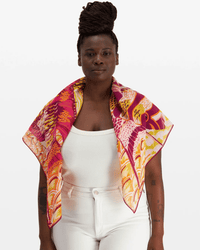 A woman with braided hair in a bun wears the Inoui Editions Square 100 Tango in Yellow, an oversized bandana, over a white sleeveless top and high-waisted white pants, standing against a plain white background.