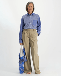 A barefoot older woman with gray hair wears a blue striped shirt and high-waisted beige trousers. She stands against a plain background, holding the Square 100 Yoga in Blue bandana by Inoui Editions in her left hand.