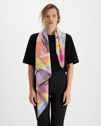 A woman in a black outfit faces forward, wearing the Square 100 Yoga in Multicolor scarf by Inoui Editions, a cotton-silk blend, draped around her neck against a plain white background.