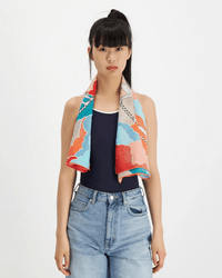 A woman with long black hair in a high ponytail wears a sleeveless navy top, light blue jeans, and the Inoui Editions Square 100 Yoga in Turquoise bandana around her neck, standing against a plain white background.