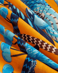 A close-up of the Square 130 Cerise in Orange by Inoui Editions—folded cotton fabric with blue and black zigzags, abstract shapes, and birds on a bright orange background—ideal for an oversized bandana.
