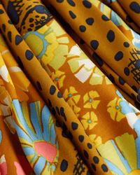 The Inoui Editions Square 130 Pampa in Caramel is a vibrant silk modal bandana with yellow, blue, and red floral and abstract patterns accented by black dots on a golden caramel background, softly draped with visible folds.