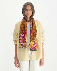 A woman with straight gray hair wears a beige shirt over a white top and pants, accented by an Inoui Editions Square 130 Pampa in Fuchsia scarf in vibrant tones, standing against a plain white background.