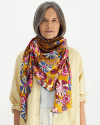 A woman with long gray hair wears a cream shirt over a white top and the Square 130 Pampa in Fuchsia scarf by Inoui Editions, featuring vibrant floral and geometric patterns, standing against a plain white background.