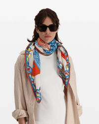 A woman wearing dark sunglasses, a white top, a beige striped shirt, and the Inoui Editions Square 130 Tango in Orange stands against a plain white background.