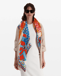 A woman in a white dress, beige striped jacket, and black sunglasses stands against a plain white background, draping the Inoui Editions Square 130 Tango in Orange oversized bandana elegantly over her shoulders.