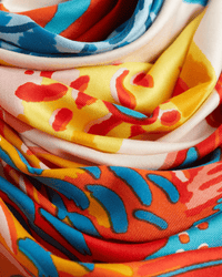 A close-up of swirling, colorful fabric folds. The Inoui Editions Square 130 Tango in Orange bandana showcases bold abstract designs in vibrant red, orange, blue, yellow, and cream—a striking statement accessory.