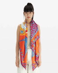 A woman with long black hair and bangs wears Inoui Editions' Square 130 Tango in Red, an oversized colorful silk-modal bandana scarf, draped over a light sleeveless top and white pants, standing against a plain white background.