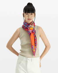 A woman with straight black hair in a high ponytail wears a sleeveless beige top, white pants, and the Square 130 Tango in Red silk modal blend bandana by Inoui Editions, standing hands-on-hips against a plain background.