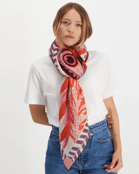 A woman in a white T-shirt and blue jeans models the Inoui Editions Square 130 Toucan in Pink, a silk cashmere scarf with bold pink, red, and purple patterns, tied loosely around her neck against a plain white background.