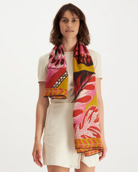 A woman in a short-sleeved cream dress stands against a plain background with the Inoui Editions Square 130 Toucan in Yellow scarf, featuring vibrant pink, red, yellow, black, and white accents, elegantly draped around her neck.