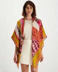 A woman in a cream dress stands facing forward, draped in the Inoui Editions Square 130 Toucan in Yellow scarf, featuring a vibrant toucan design with pink, red, and yellow hues against a plain background.