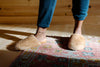 Person wearing tan, fluffy slippers and blue pants, standing on a colorful, patterned rug in a warmly lit room.