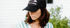 Person wearing a black cap and a striped shirt, looking downward. The background features a white wall and green foliage.