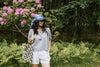 Woman standing outdoors in a garden with pink flowers, wearing a blue hat, gray short-sleeve top, white shorts, and holding a patterned tote bag.