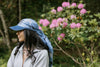 A woman with long dark hair wearing a blue scarf and cap stands outdoors near a shrub with clusters of pink flowers.