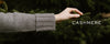 A hand in a grey cashmere sleeve holds a pine branch. The word 