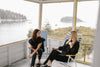 Two women sitting in striped chairs on a screened porch overlooking a lake with a forested background.