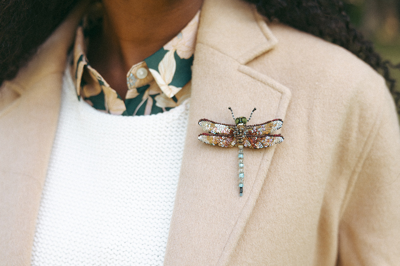 A person wearing a beige blazer with a large, intricately detailed dragonfly brooch pinned to the lapel.