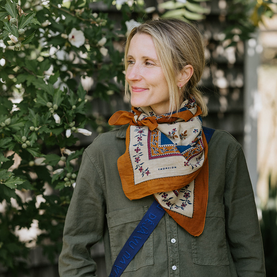 Person wearing a knit sweater and jeans holds a green woven bag with a red patterned scarf tied to it, standing outdoors.
