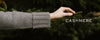 A person's arm in a gray cashmere sweater reaching out to touch a green fir tree branch, with the word 