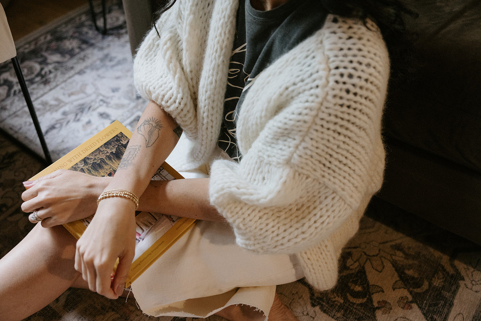 A woman wearing a striped cardigan stands near trees and a body of water, smiling and looking down. Autumn leaves surround her.
