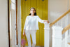 A person stands by a staircase, wearing a white top and pants, and holding a multicolored bag. The door behind is yellow.